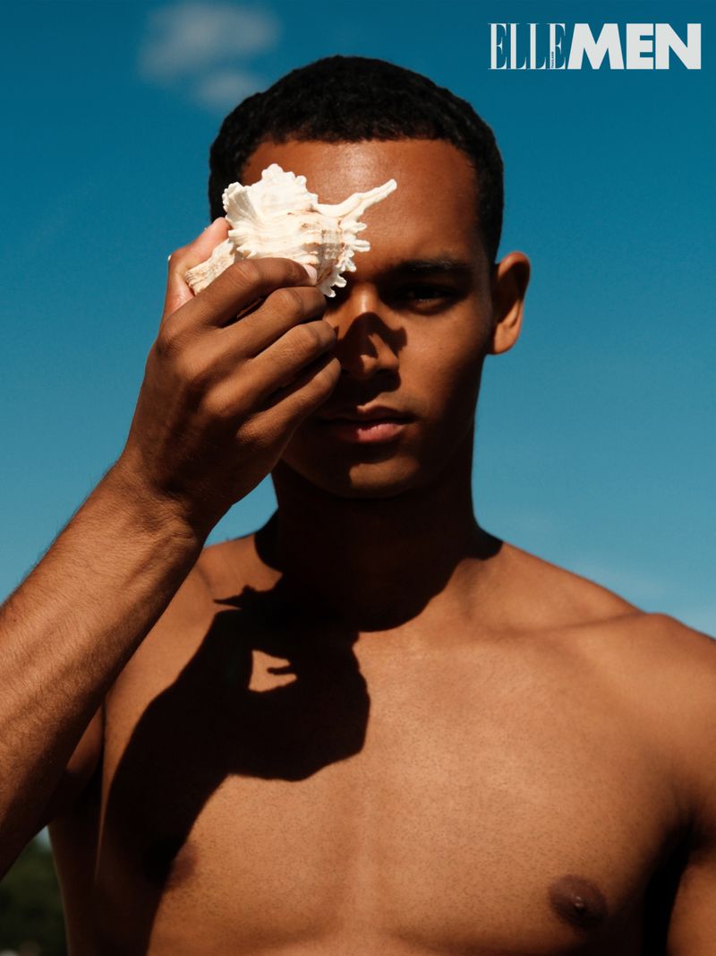 Portrait of dark-skinned man holding seashell to face against clear blue sky; shirtless male fashion shot for Elle Men's editorial content.