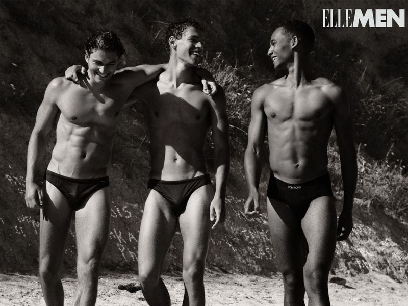 Black and white shot of three adult men standing outside, wearing swim briefs. They are embracing, with greenery in the background.