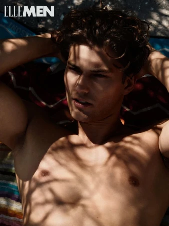 A tan, shirtless adult male model poses with hands behind his head, laying on a multicolored surface. Sunlight creates leaf shadows on his body.