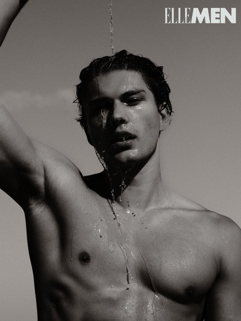 Monochrome portrait of a shirtless male model with water pouring over his head and streaming down his face against a clear sky background.