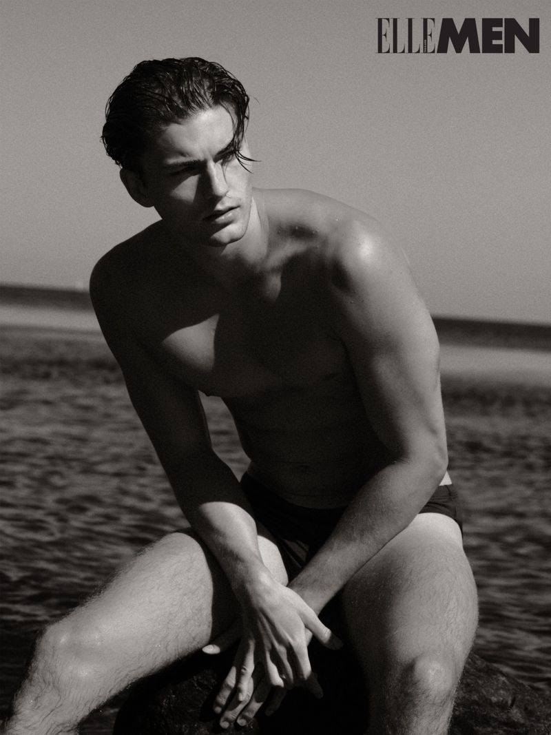 A shirtless, athletic adult male model sits on a rock in ocean water with the Elle Men magazine logo in the upper-right corner in a black and white image.