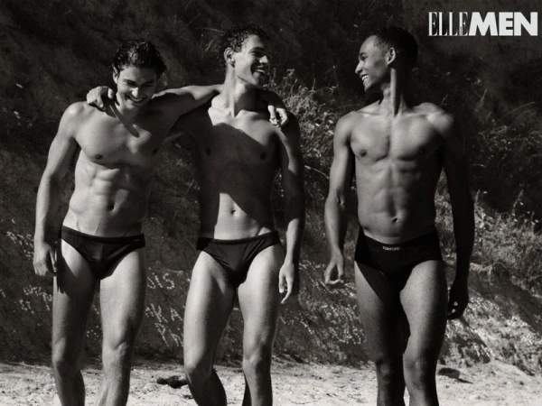 Three male models in black swim briefs walking and smiling together outdoors against a natural rock background