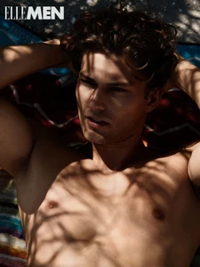 Shirtless man with brown curly hair lying down on a towel with dappled tree shadows across his face and chest.