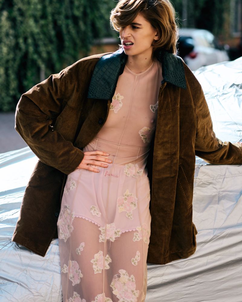 Adult female model wearing a sheer floral dress and brown corduroy jacket posing outside with a blurred background of trees and cars.