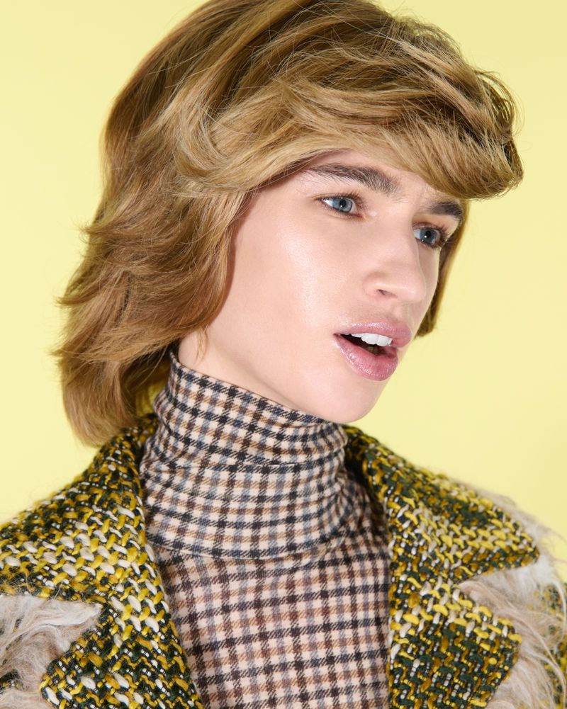 Close-up studio shot of a young adult with blonde wavy hair, blue eyes, wearing a brown and black checkered top, and a green tweed jacket.