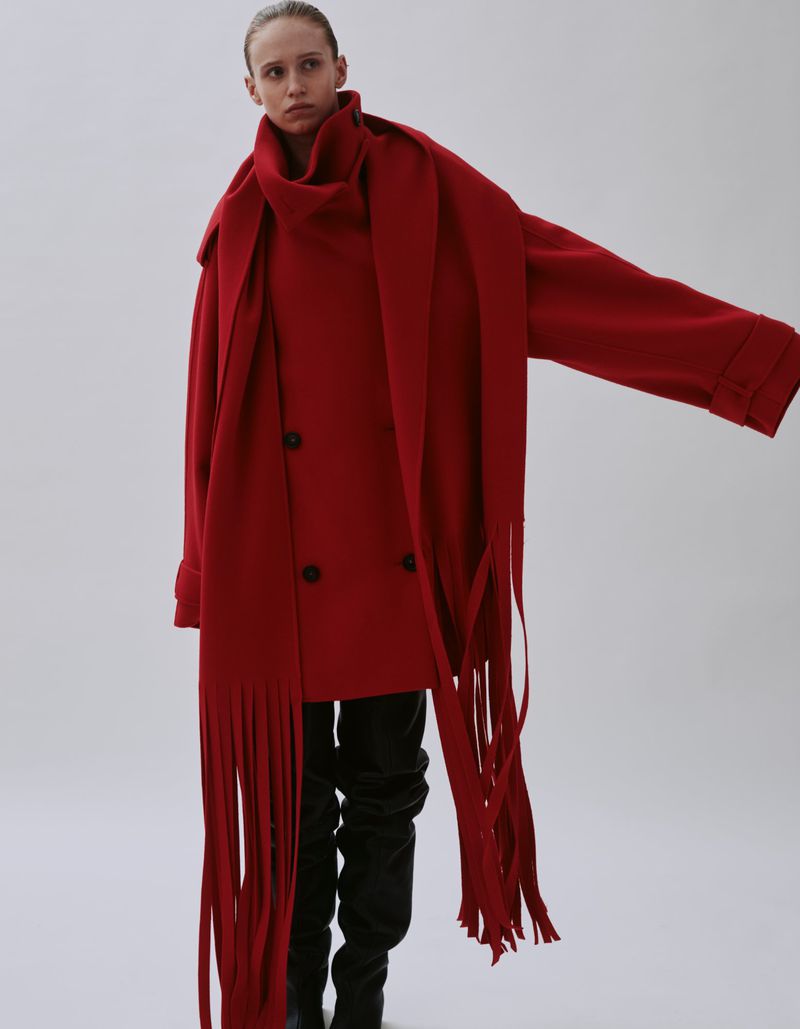 A woman in a red double-breasted coat and matching fringed scarf poses in front of a white background, showcasing a fashion-forward winter outfit.