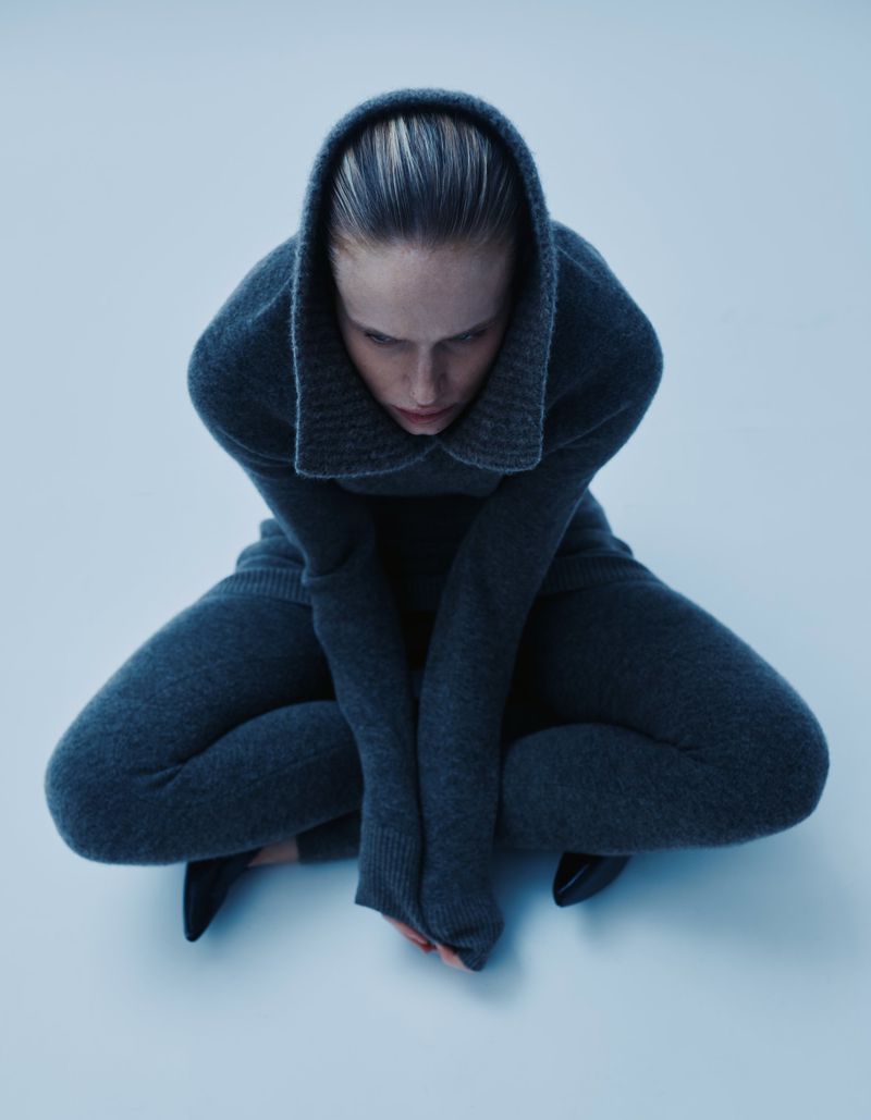 A woman in a grey hooded sweater and pants sits in a low squat on a pale blue background looking down in a high-angle studio shot.
