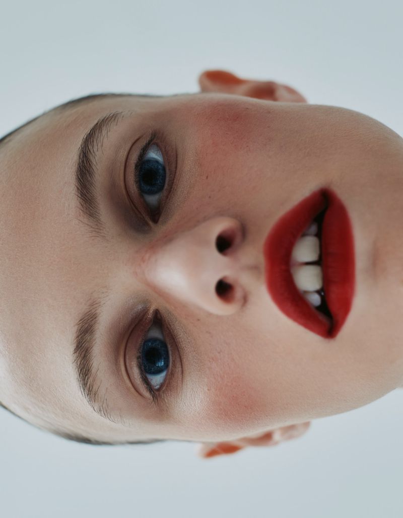 Close-up portrait of a woman with red lipstick and blue eyes against a light grey background, beauty and fashion headshot.