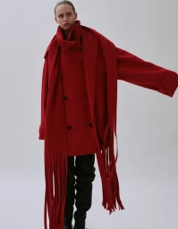Model wearing a structured red coat with a long fringed scarf and black boots against a neutral grey studio background