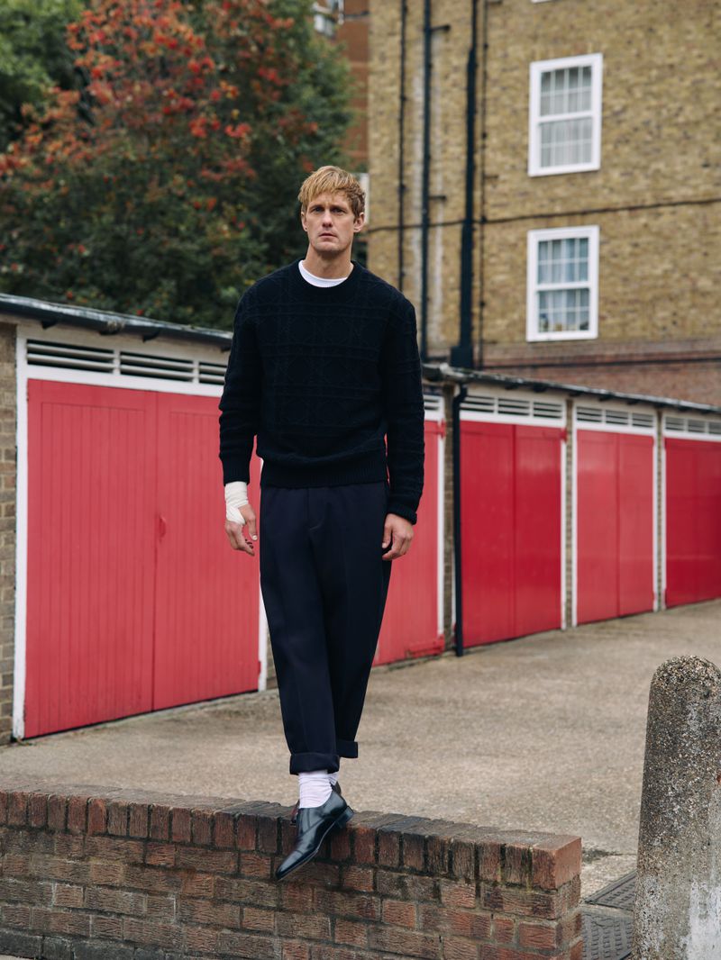 Blond male model wears dark sweater and trousers while walking on a wall outside, featuring red garage doors and brick building.