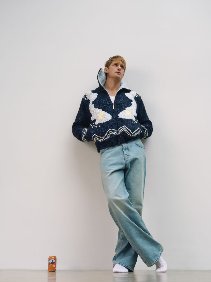 Young man wearing a navy sweater and light blue jeans leans against a white wall with an orange can on the floor next to him.