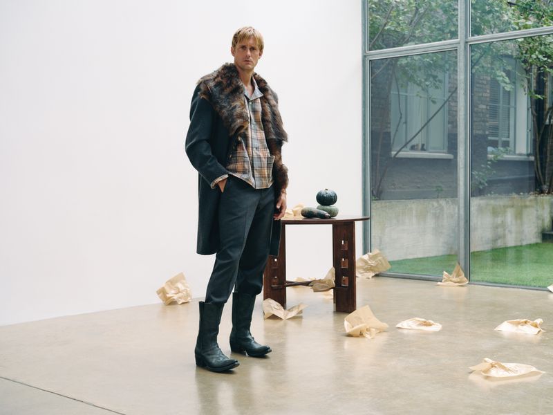 A man stands in a modern interior with a fur-collared coat, plaid shirt, and boots next to a minimalist table with squash in a minimalist setting.