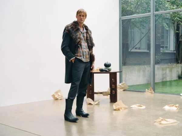 A man stands in a modern interior with a fur-collared coat, plaid shirt, and boots next to a minimalist table with squash in a minimalist setting.