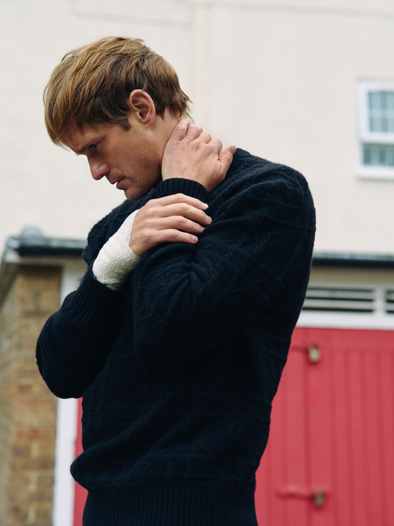 Blond male model in a black sweater stands outdoors with bandaged wrists against a red door and a white building, moody and contemplative.