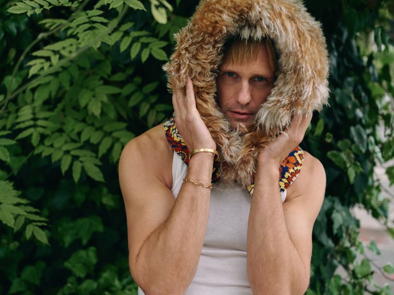 Portrait of a fair-skinned adult male in a white tank top and fur-lined hood with green foliage in the blurred background.