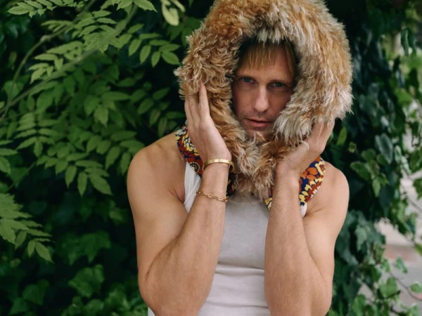 Portrait of a fair-skinned adult male in a white tank top and fur-lined hood with green foliage in the blurred background.