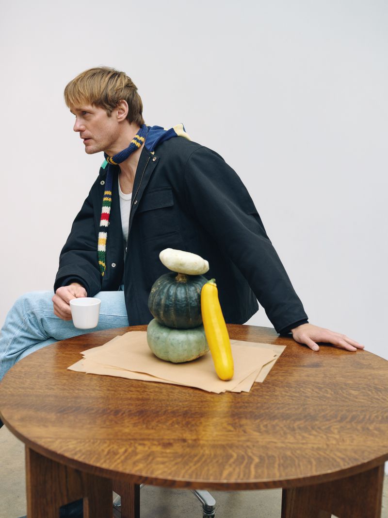 Adult male in a black jacket seated near a round wooden table with a stack of butternut squash and zucchini, studio lighting.