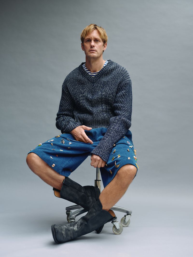 Adult male seated on a rolling chair in a photo studio wearing a sweater, patterned shorts, and boots against a grey background.