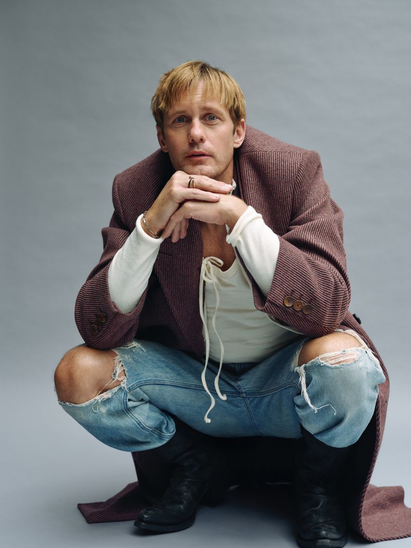 Man with blond hair in a long brown coat, ripped jeans and black boots squatting in front of a grey background looking at the camera.