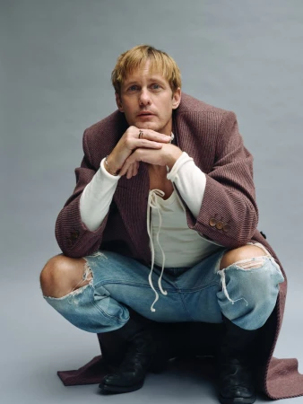 Man with blond hair in a long brown coat, ripped jeans and black boots squatting in front of a grey background looking at the camera.