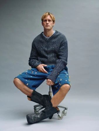 Alexander Skarsgård seated on a wheeled stool wearing a dark knit sweater, blue textured shorts, and black boots against a grey backdrop