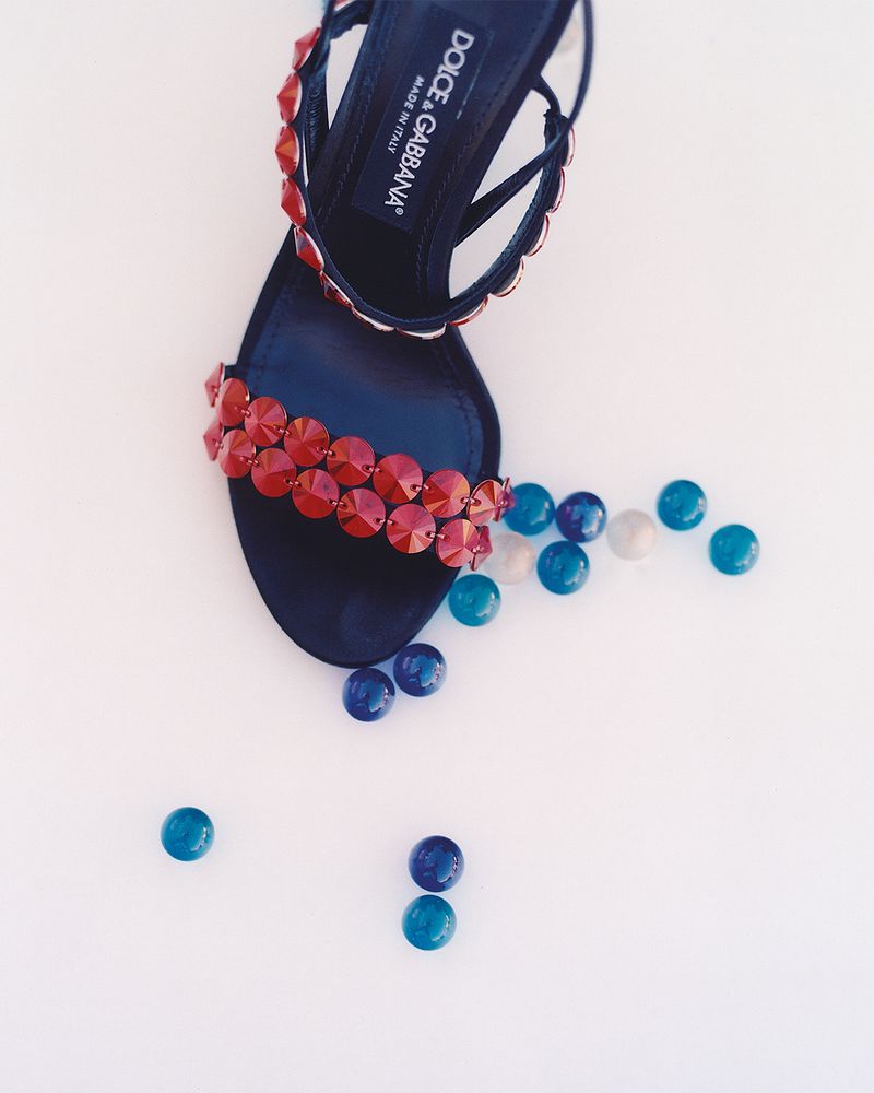 Fashion still life featuring a high-heeled Dolce & Gabbana sandal with red bejeweled straps, resting on a white surface and accented with blue glass marbles.