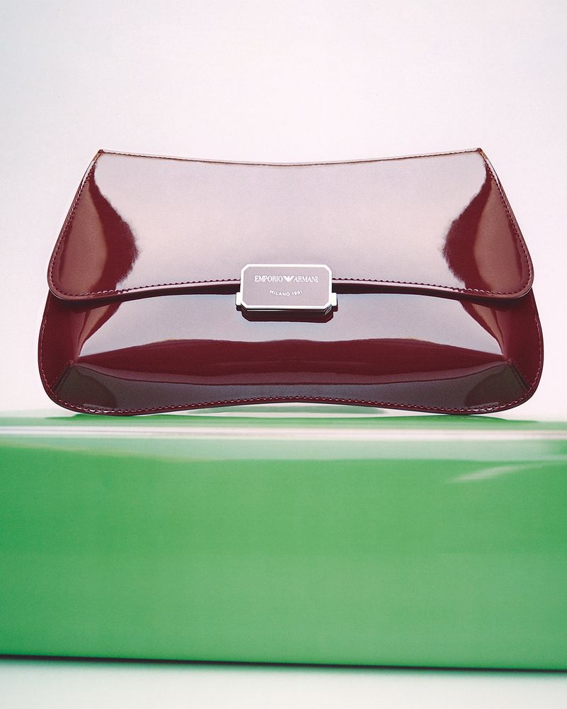 Close-up studio shot of a shiny burgundy clutch purse with a silver clasp and brand logo, displayed on a glossy green surface.