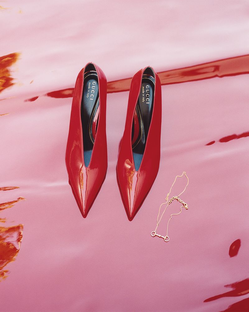 Overhead shot of a pair of red patent leather high heels on a pink shiny surface with a gold necklace, capturing a modern fashion aesthetic.