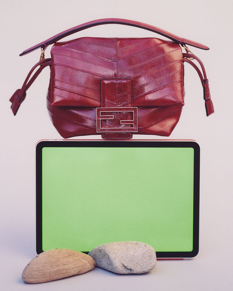 Studio shot of a red leather handbag with metal accents displayed on a tablet computer with stones, against a neutral backdrop.