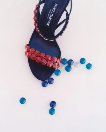 Overhead studio shot of a black high-heeled sandal with red crystal embellishments on a white background with blue beads.