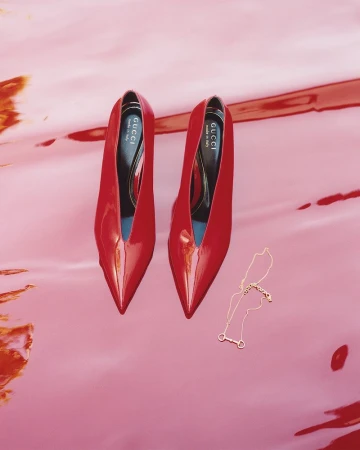 Glossy red pointed-toe high heels placed on a glossy red surface next to a delicate gold necklace