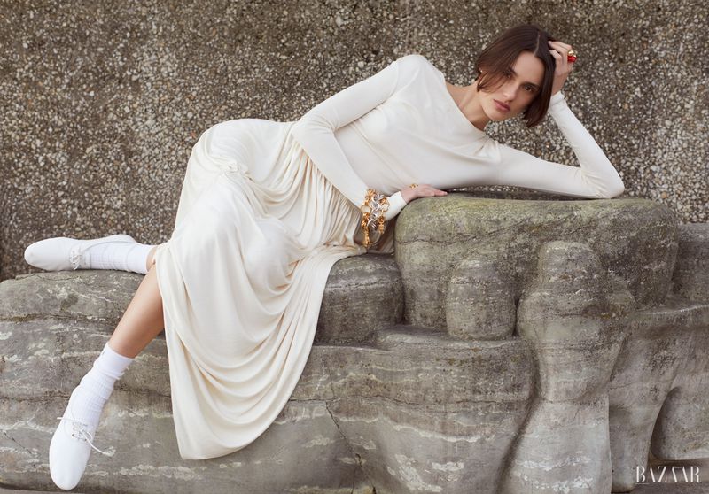 A woman in a long white dress poses on a rock, showcasing fashion in an outdoor setting with soft, diffused lighting.