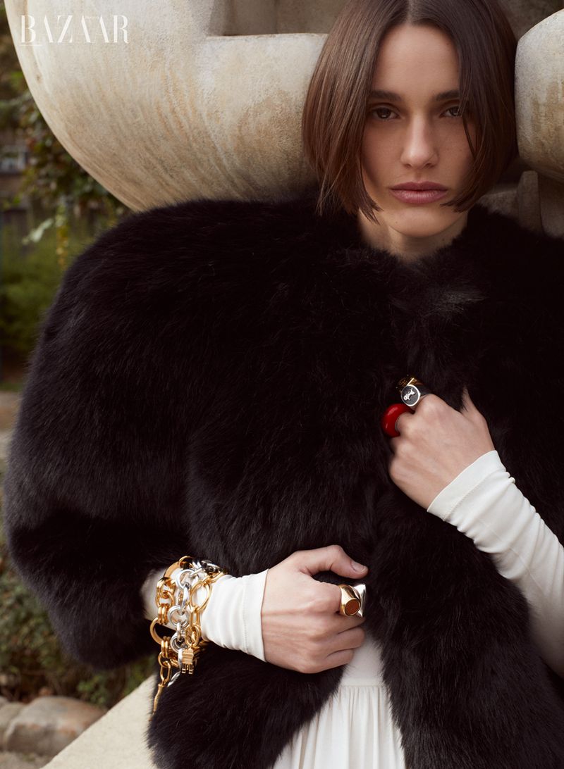 Fashion editorial shot of a woman wearing a black fur coat, jewelry, and a white dress, posing in front of an abstract stone sculpture.
