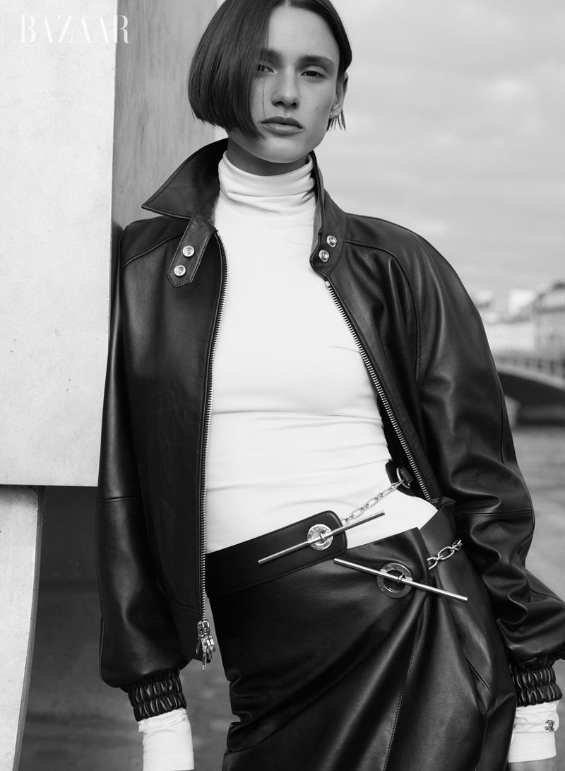 Black-and-white fashion portrait of a woman in a leather jacket and skirt leaning against a wall with a city scene in the background.