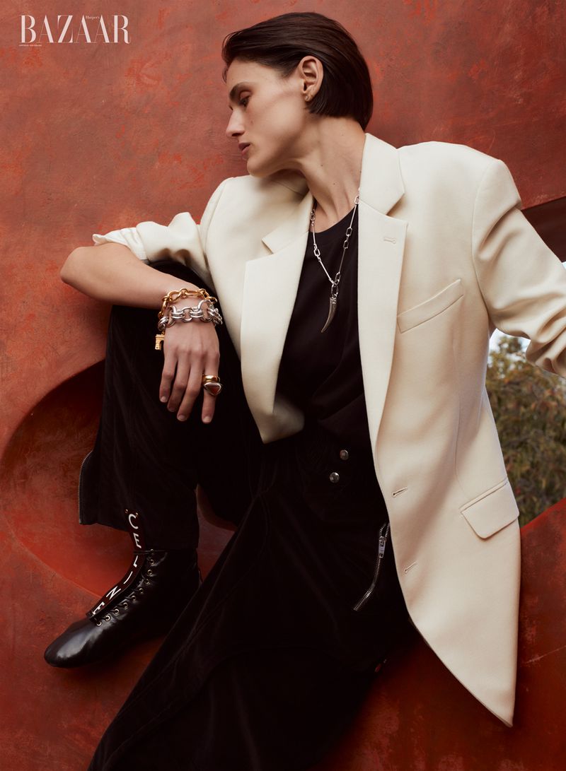Fashion editorial shot of an adult woman in a cream blazer, black pants, and lace-up boots, posed against a copper metal backdrop.