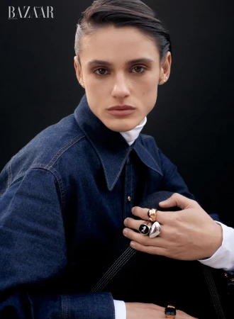 Portrait of a woman with slicked-back dark hair wearing a denim jacket, white turtleneck, and rings, posing against a dark background.