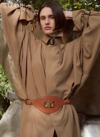 Model Aleyna Fitzgerald wearing a camel coat and tan belt with arms raised outdoors for Harper's Bazaar Australia
