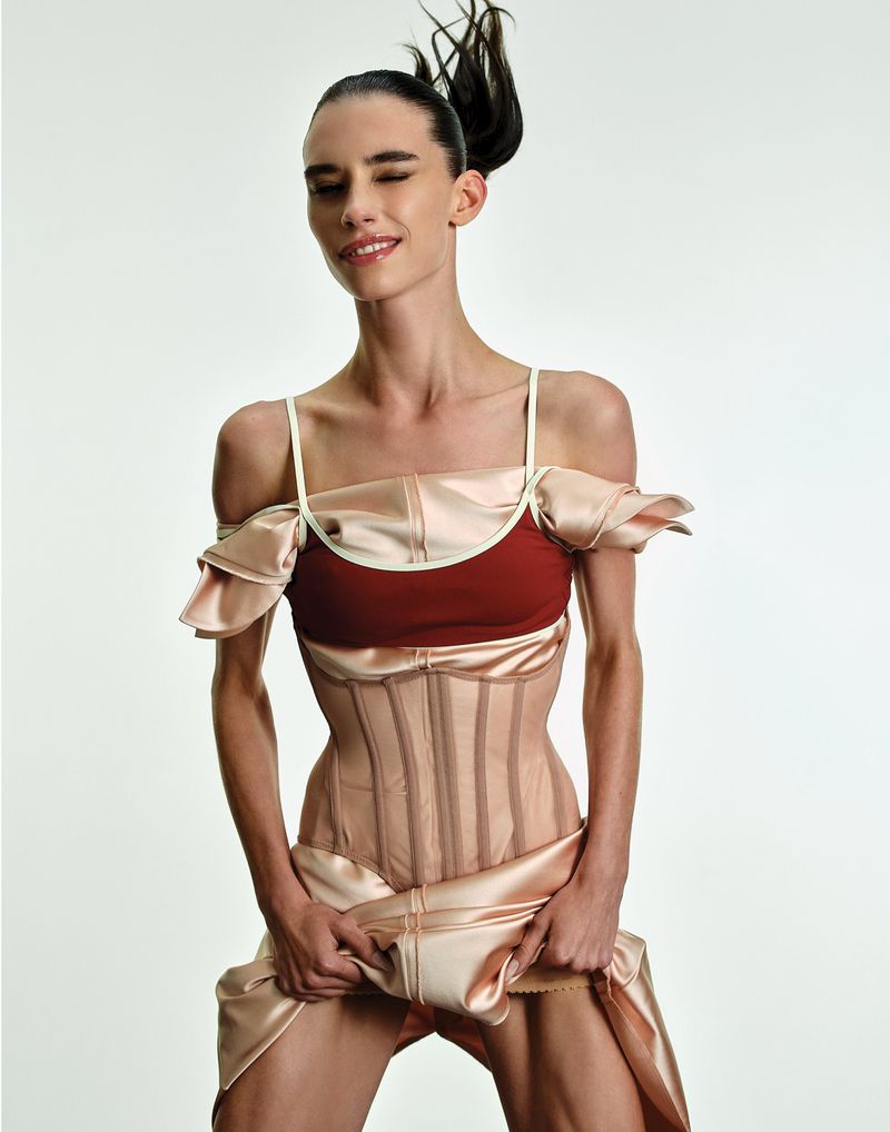A young adult woman with dark hair poses in a studio wearing a corset and satin dress with a white backdrop. Smiling expression.