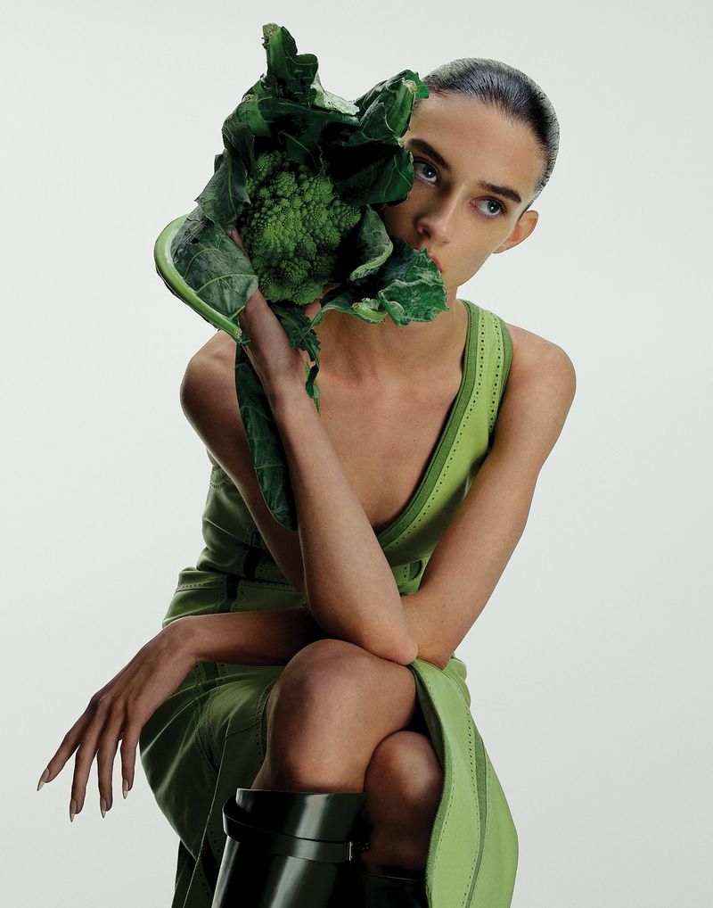 Young woman in a green dress poses in front of a white background, holding leafy romanesco broccoli covering part of her face.