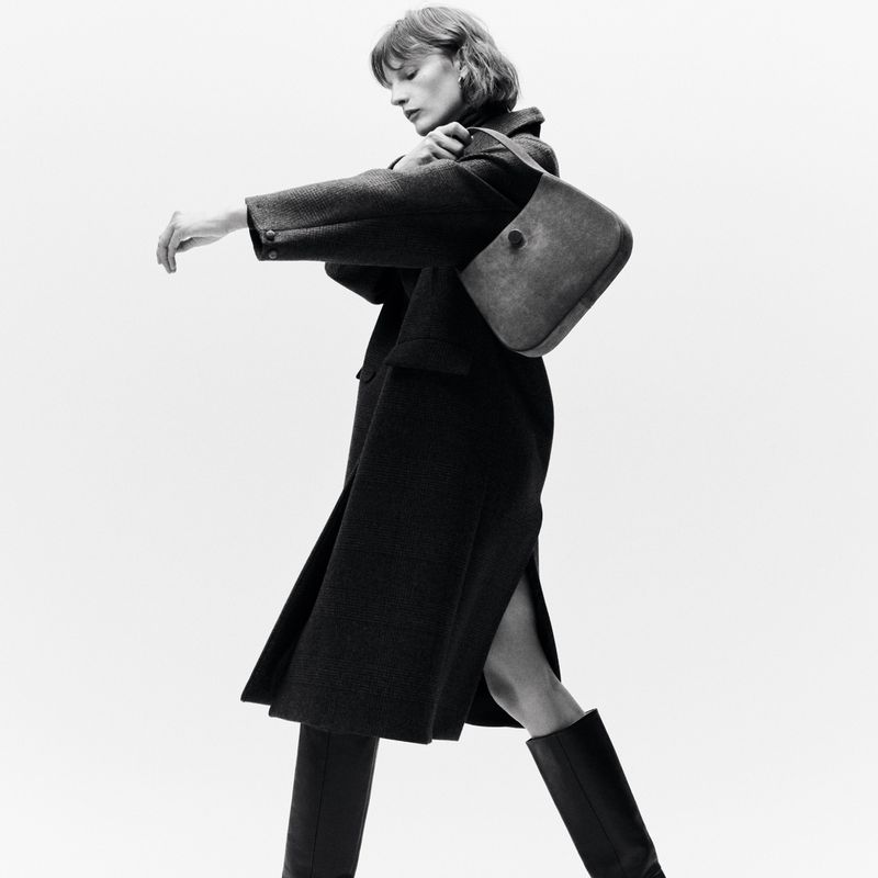 Model Sara Blomqvist wearing a dark coat and boots while carrying a shoulder bag against a white studio background.