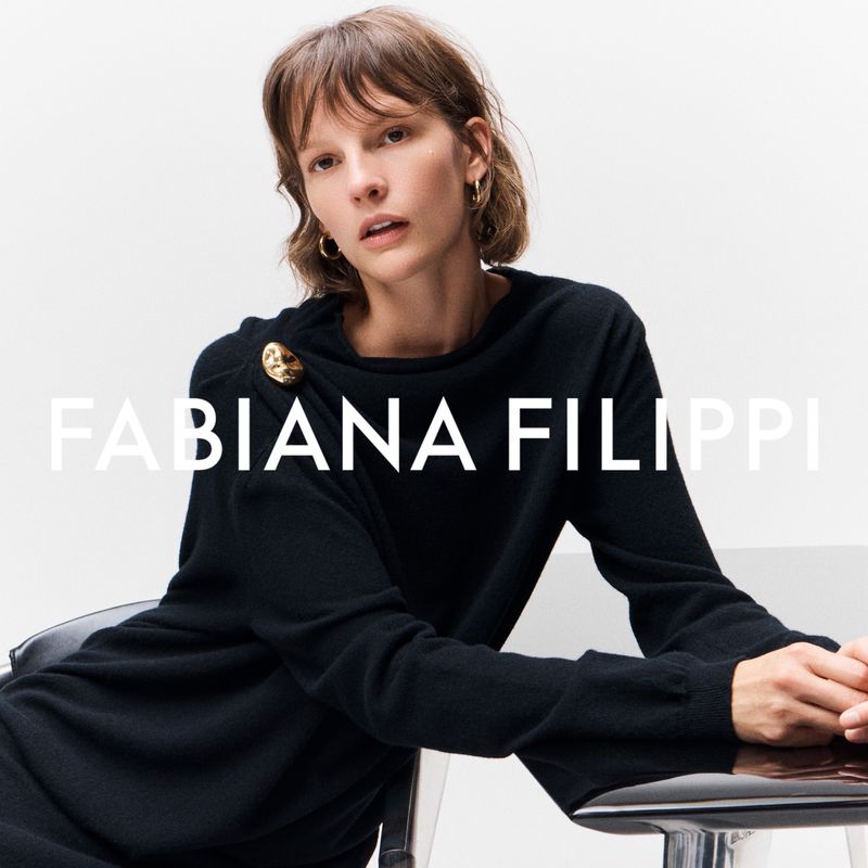 A woman models a black long-sleeved sweater with a large gold button detail against a bright white backdrop in a fashion advertisement.