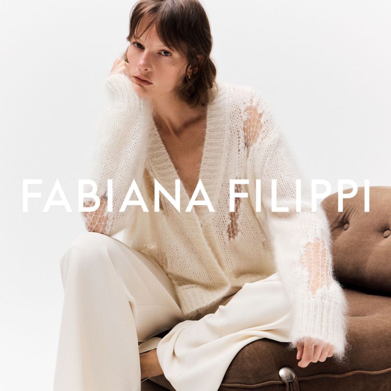 Fashion shoot of a female model in a white, distressed cardigan and pants, seated on a brown couch against a bright background.