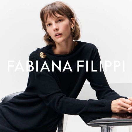 Model Sara Blomqvist seated in a black long-sleeve knit top with a gold shoulder brooch against a white background.