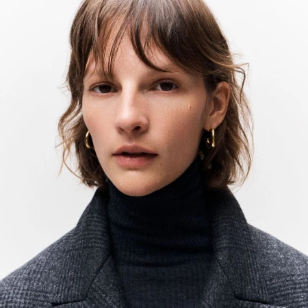 Close-up portrait of model Sara Blomqvist wearing a dark turtleneck and textured grey jacket against a white background.