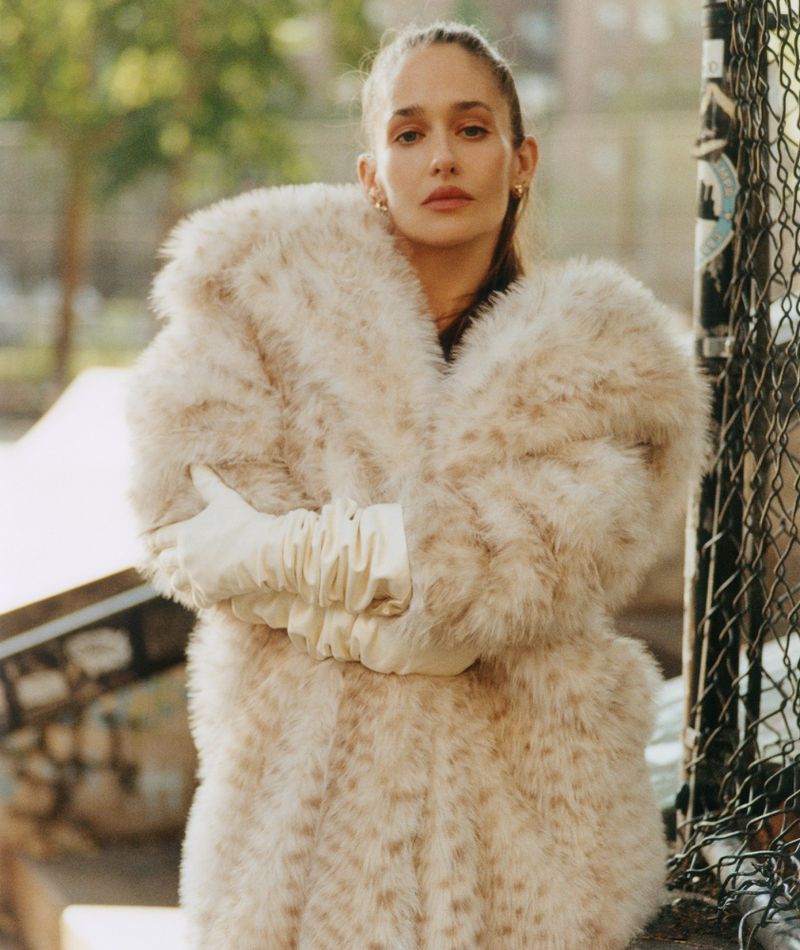 A woman posing outdoors wearing a large, fluffy faux-fur coat and white gloves, standing in front of a chain-link fence.
