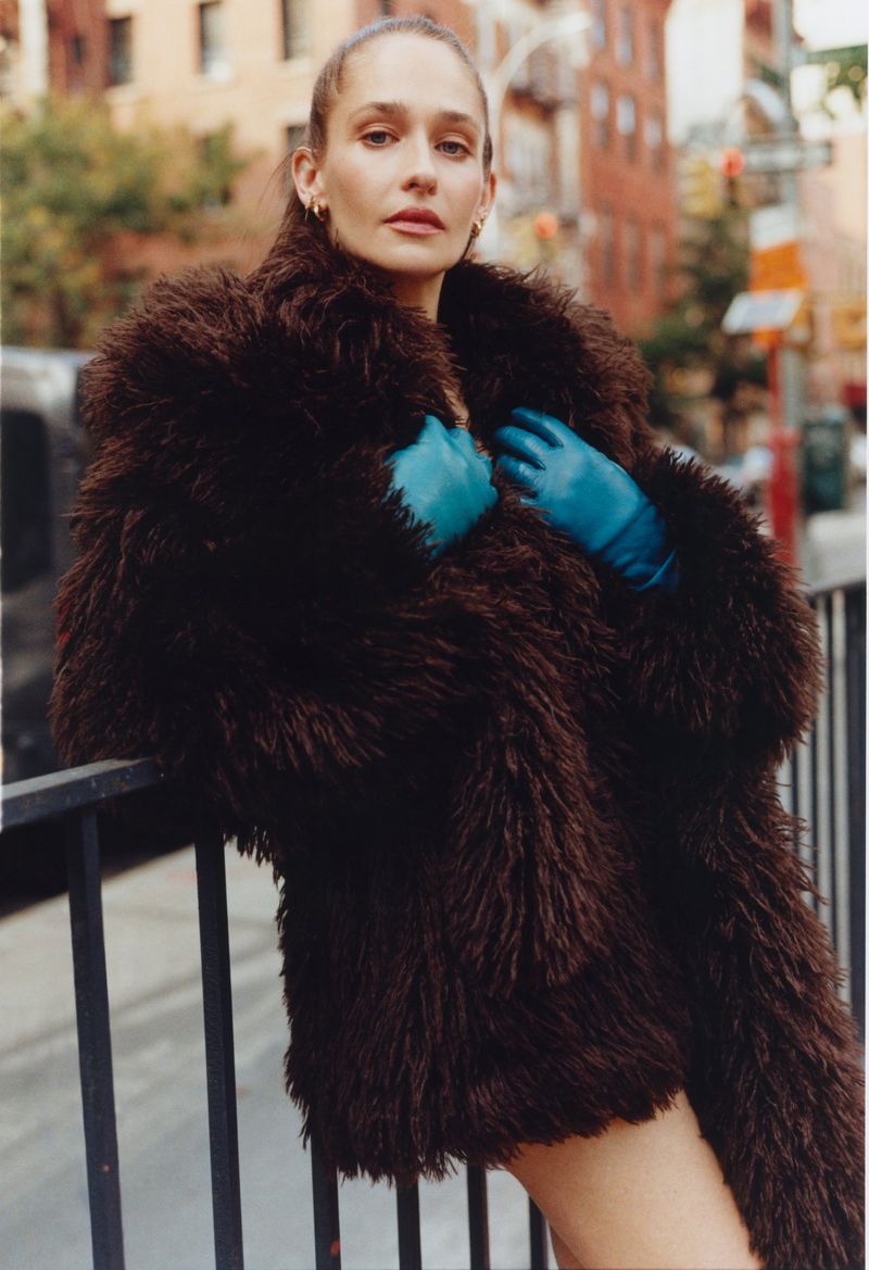 Jemima Kirke wearing a dark brown faux-fur coat and blue gloves, leaning against a railing on a city street.