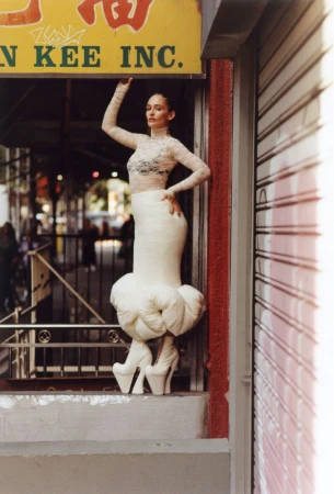 Woman poses in a lace top and avant-garde white dress and heels in front of a store with Chinese signage in an urban setting.