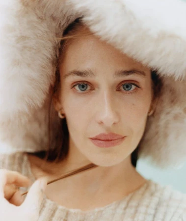 Close-up portrait of a light-skinned woman with blue eyes wearing a fur hat, looking at the camera in soft, diffused lighting.