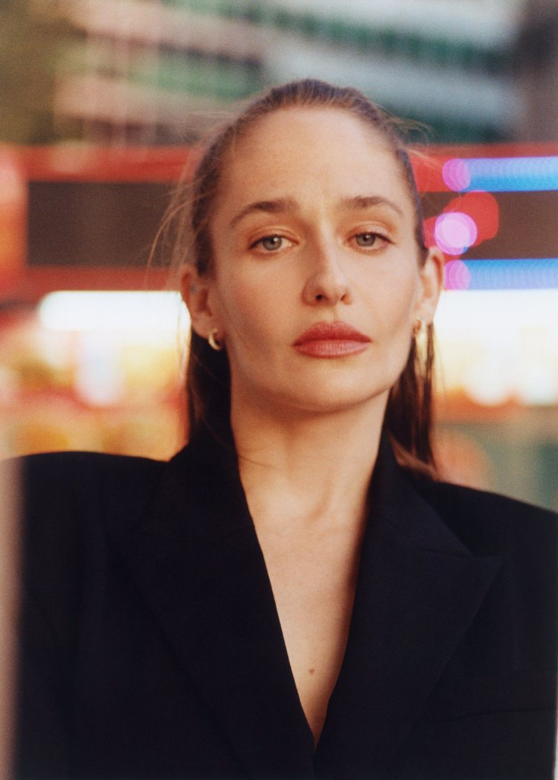 Portrait of a woman with neutral expression wearing black blazer in an urban setting with blurred neon lights at night.