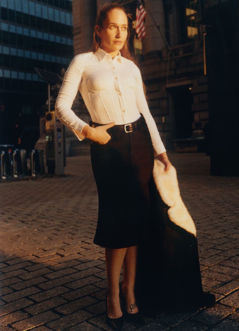 Elegant woman in white blouse, black skirt, and heels poses confidently on a brick sidewalk near buildings and an American flag.